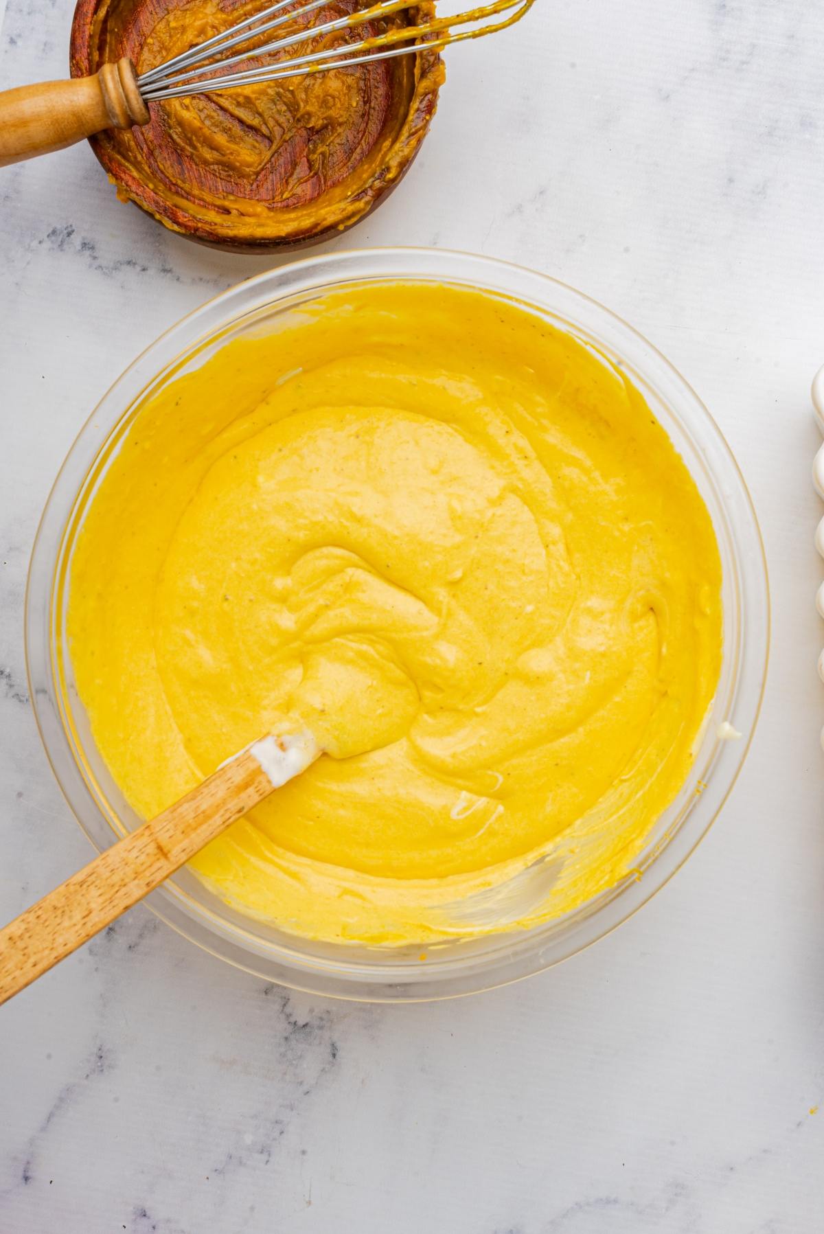 A bowl of yellow cake batter with a spatula, next to a whisk and small bowl, on a white surface.