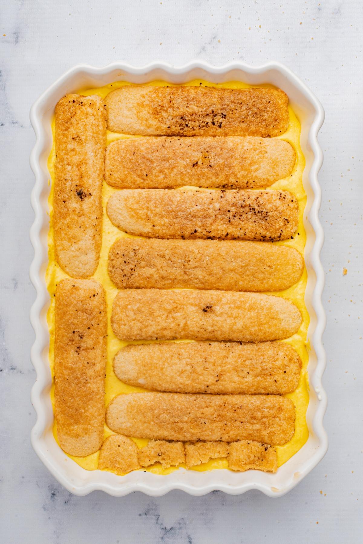 Ladyfingers layered on creamy yellow custard in a rectangular white baking dish.