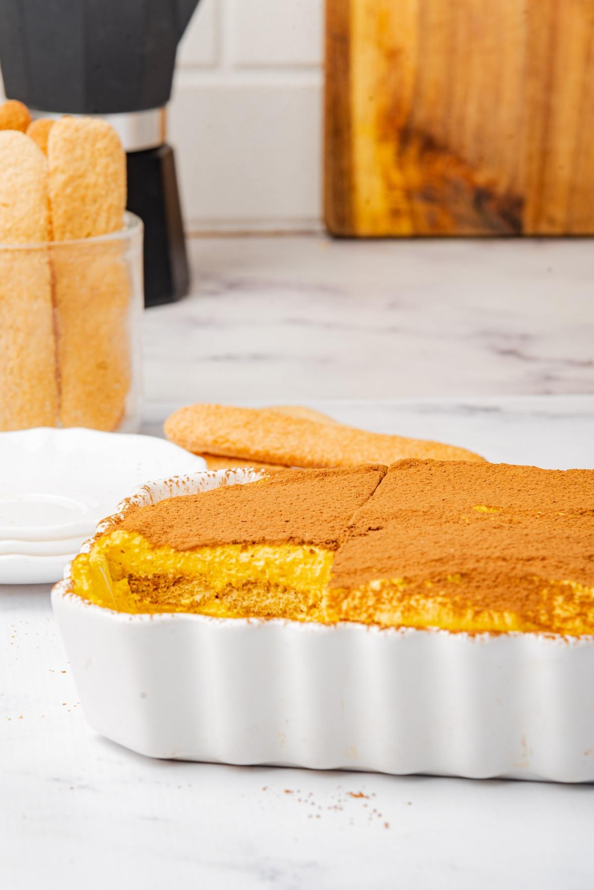 A dish of tiramisu with a slice missing, dusted with cocoa powder, and ladyfingers in the background.
