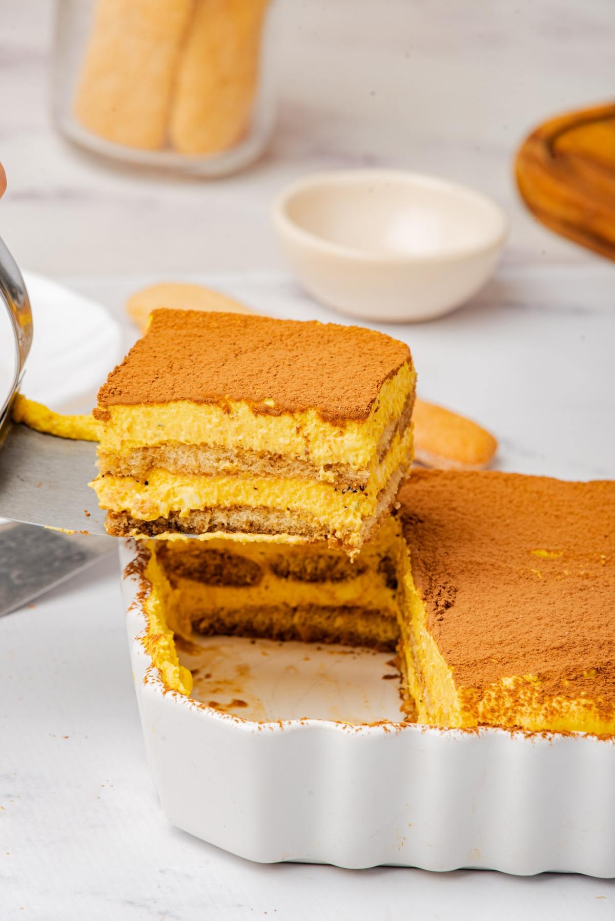 A slice of tiramisu being lifted from a white dish, showing its creamy layers and cocoa powder topping.