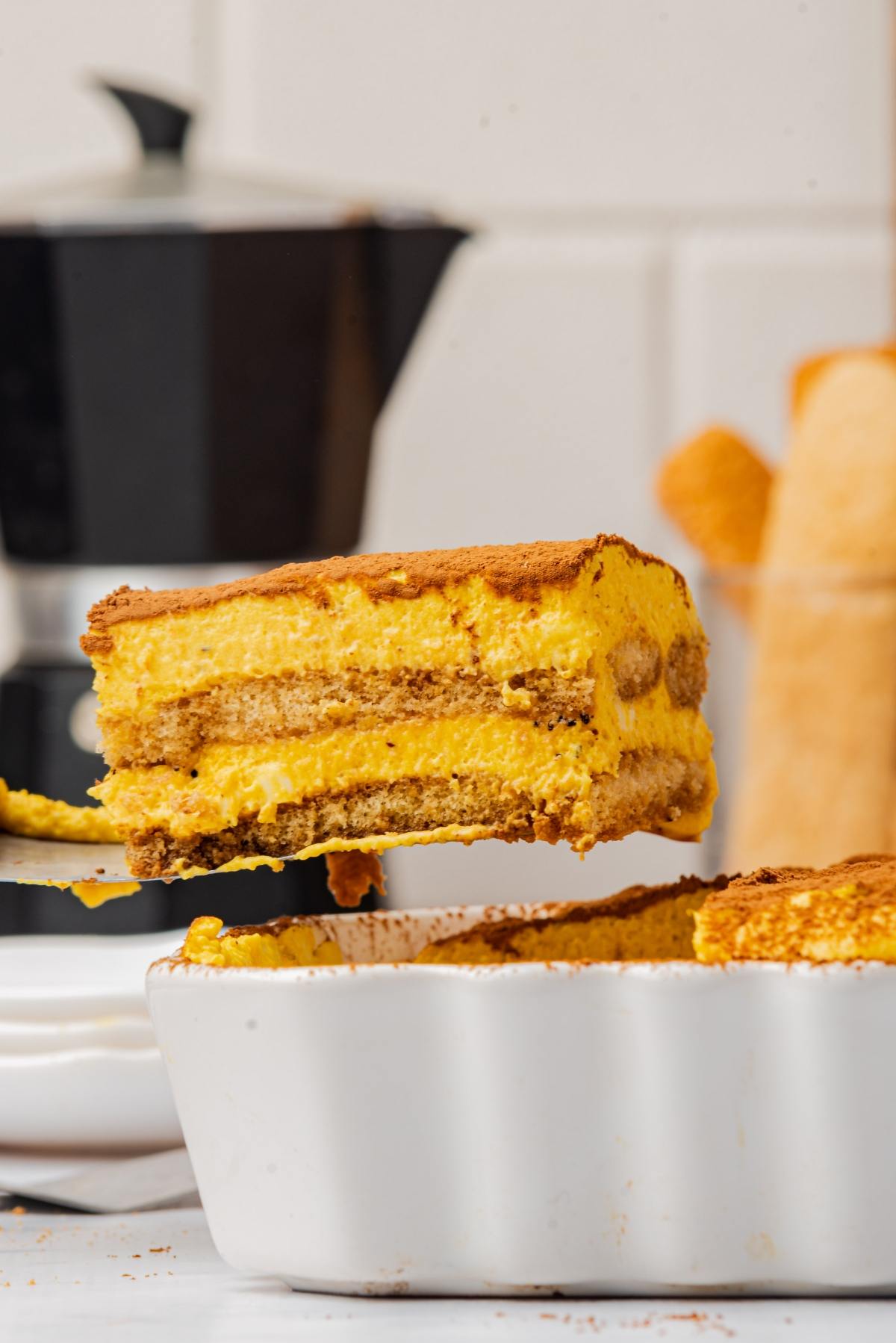 A slice of tiramisu being lifted from a white dish, with a coffee maker in the background.
