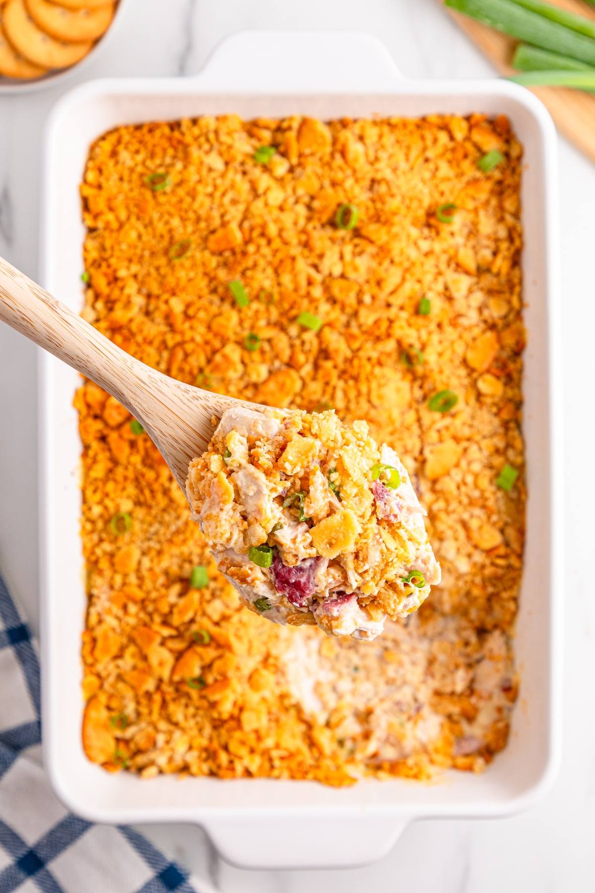 A wooden spoon holds cheesy casserole with a crunchy topping over a white baking dish of the same casserole.