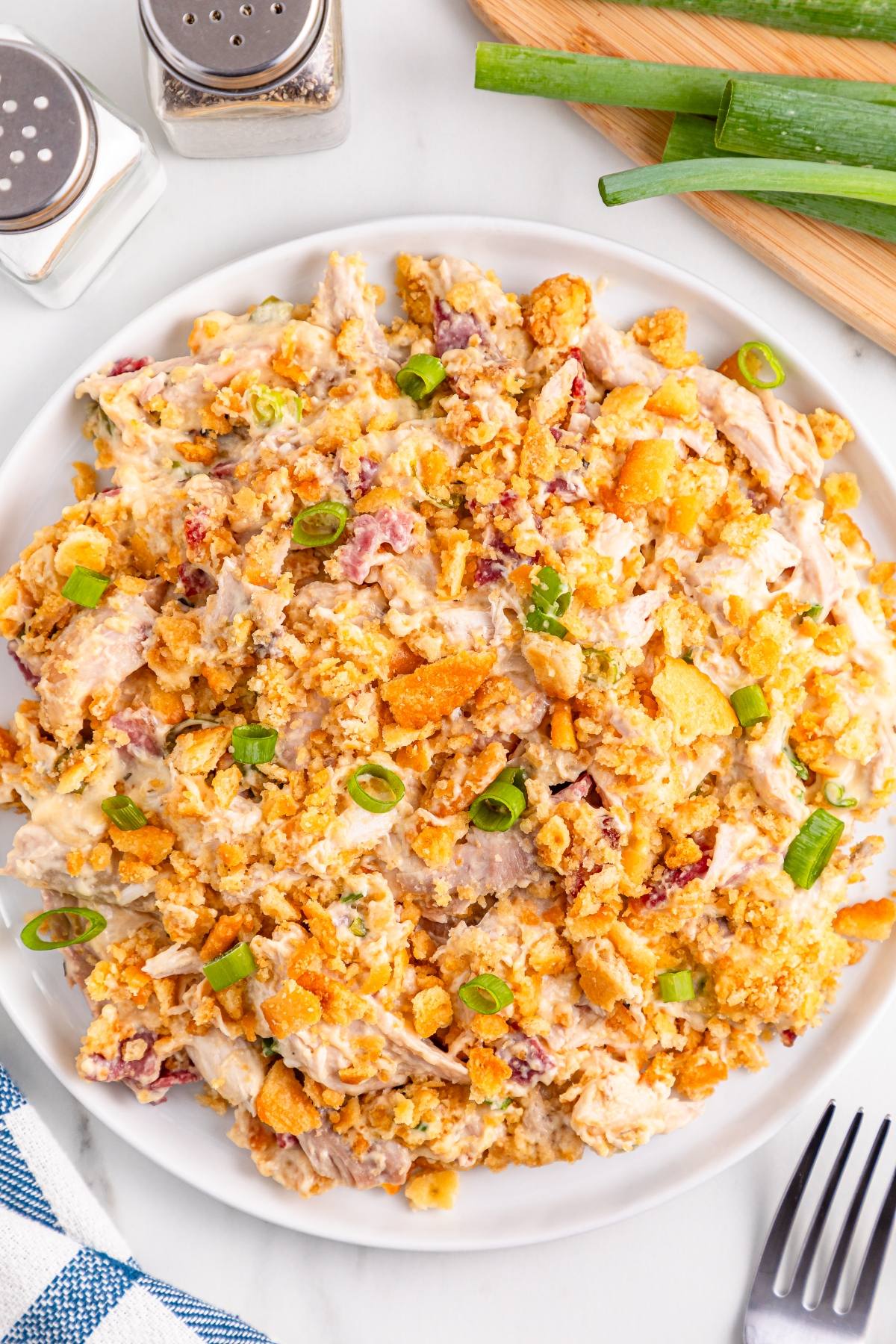 Plate of cheesy chicken casserole topped with green onions, next to salt, pepper, and a cutting board with scallions.