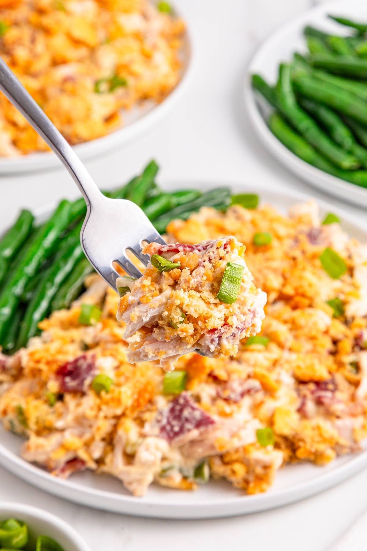A fork holds a bite of cheesy casserole with green onions above a plate with green beans and more casserole.