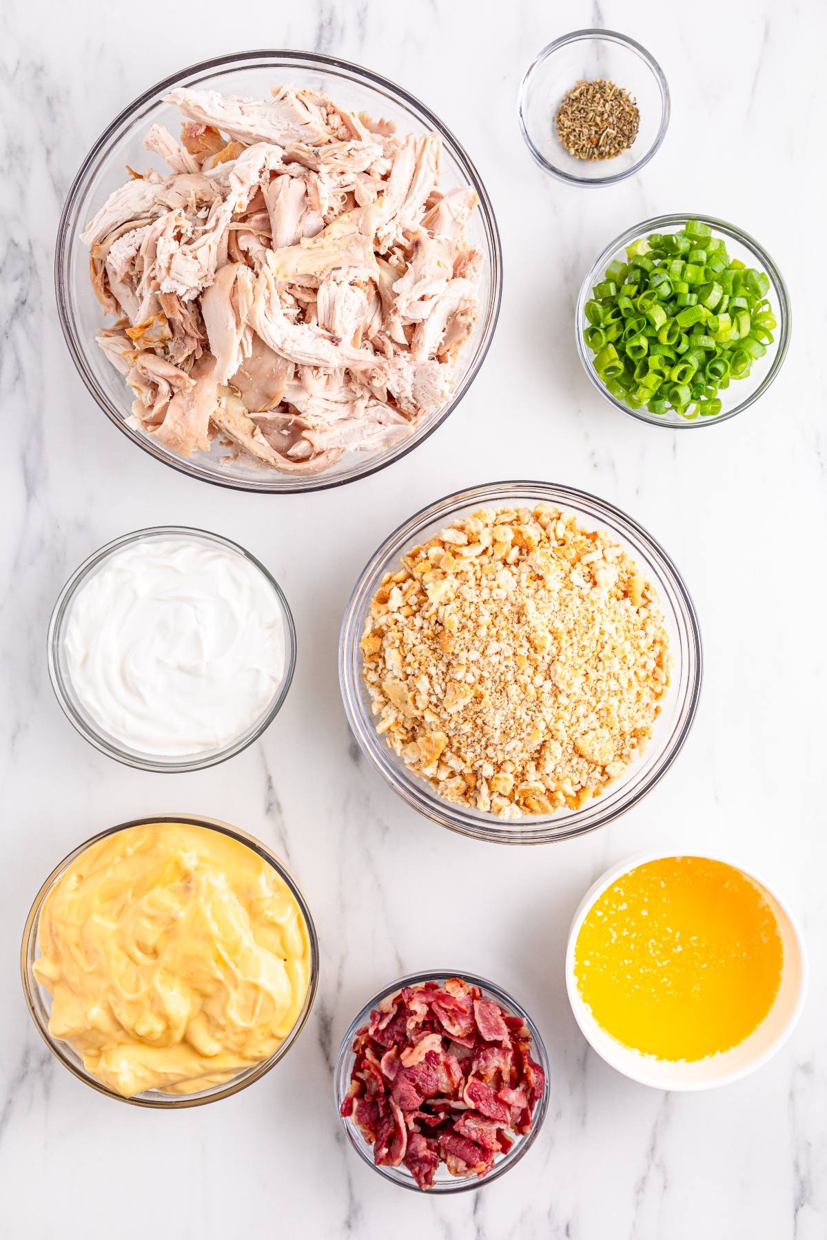 Seven bowls with shredded chicken, sour cream, crushed crackers, melted butter, cheese sauce, bacon, and green onions.