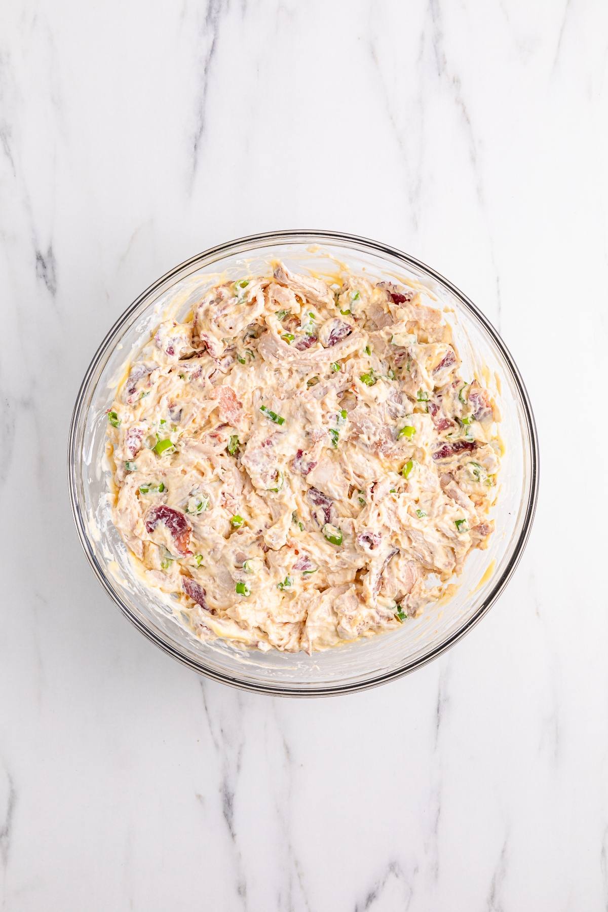 A glass bowl of creamy dip with chopped green onions and pieces of bacon on a marble surface.