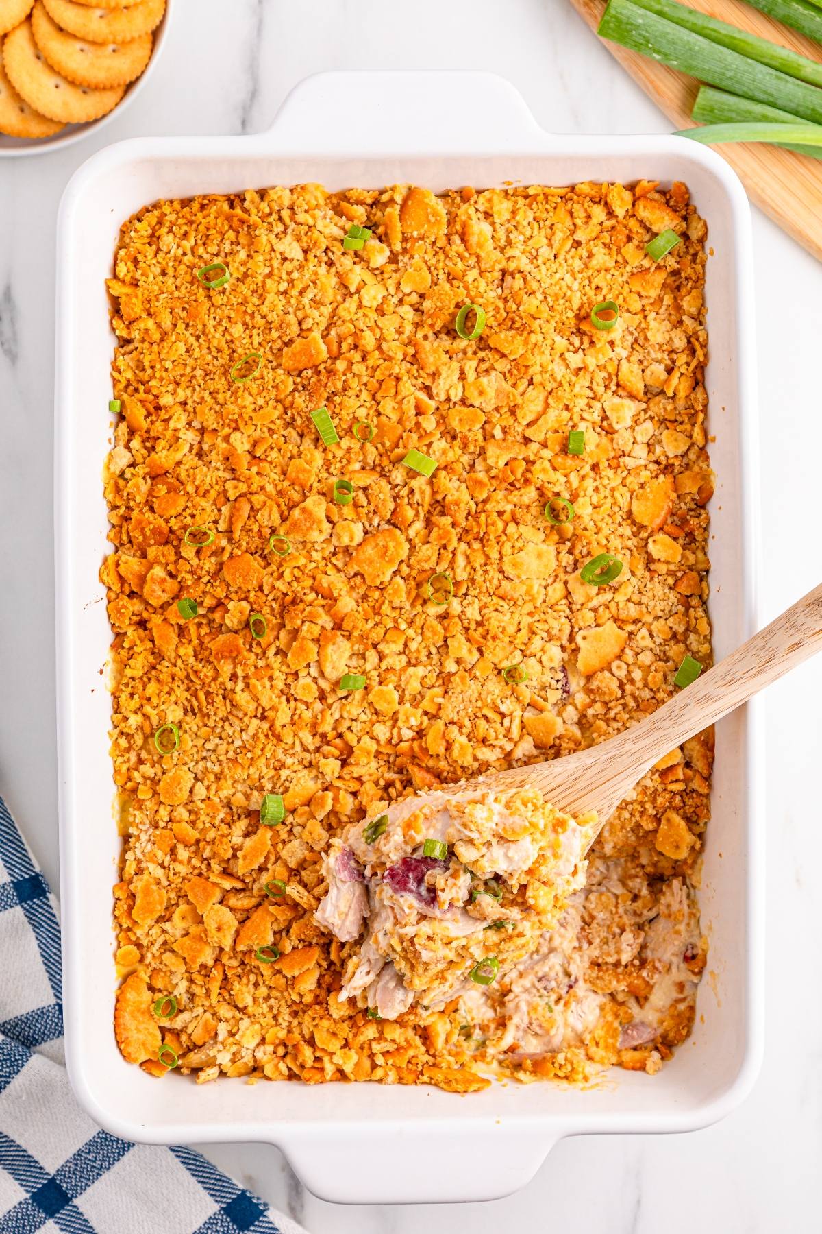 A baked casserole with a golden crumb topping, served in a white dish with a wooden spoon scooping some out.