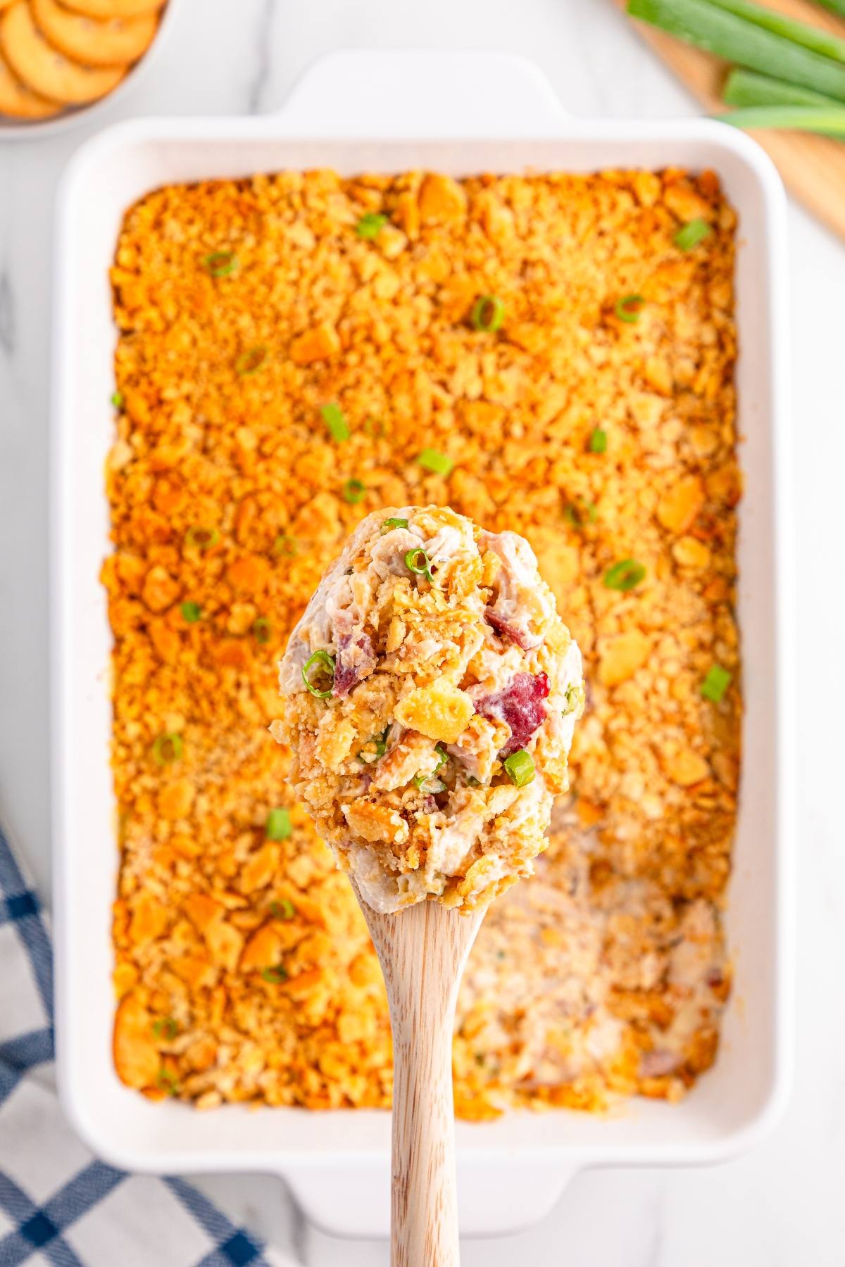 A wooden spoon holds a creamy casserole with a crunchy topping above a casserole dish.