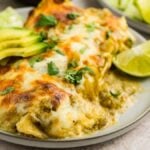 Plate of cheesy Salsa Verde Chicken Enchiladas with sliced avocado, lettuce, lime wedge, and cilantro garnish.