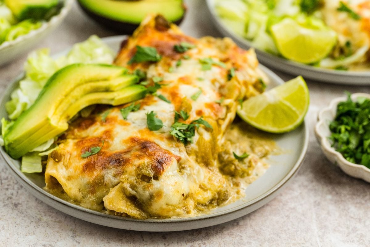 A plate of Salsa Verde Chicken Enchiladas topped with avocado slices, lettuce, a lime wedge, and sprinkled cilantro for a fresh and flavorful meal.