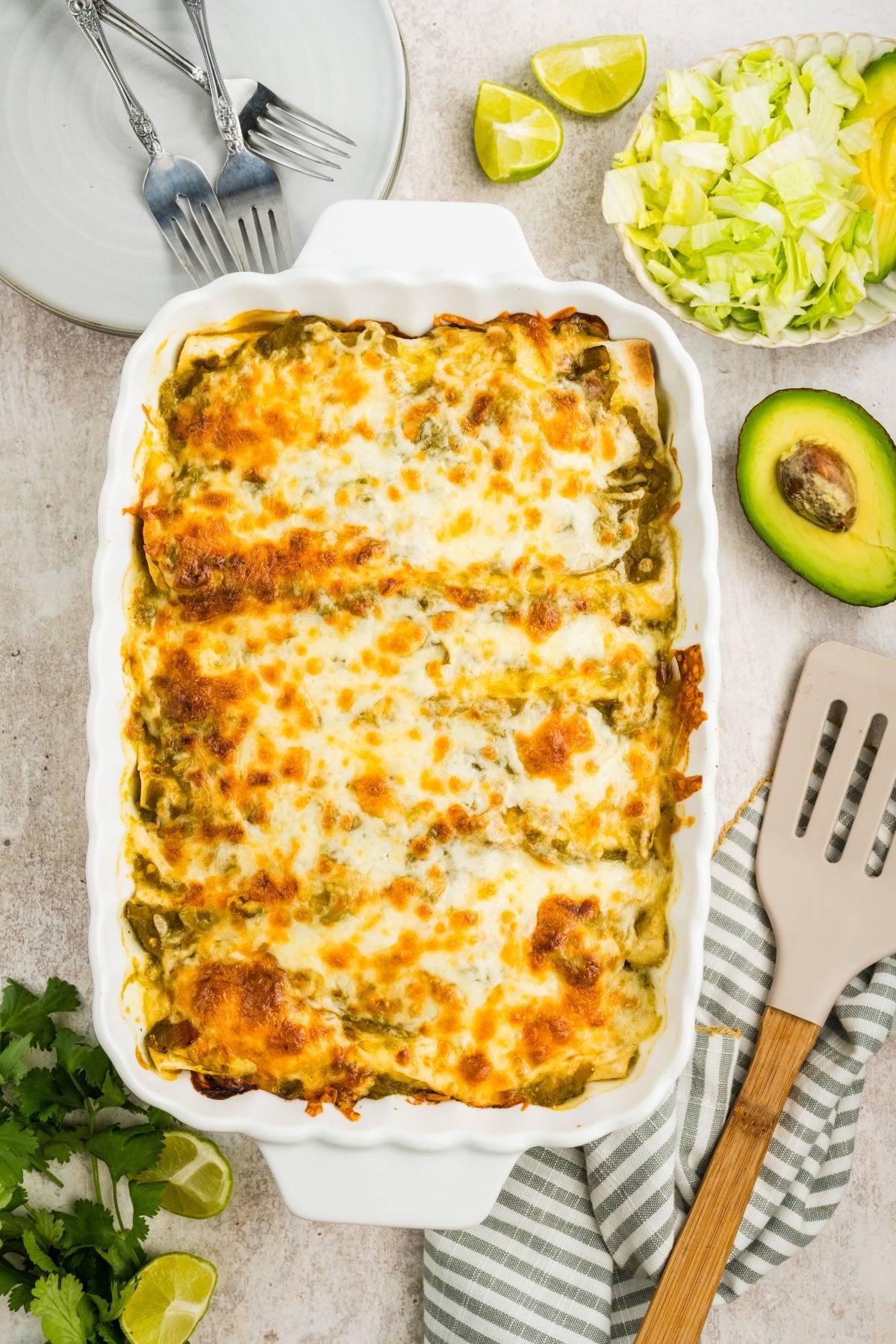 A baked casserole with melted cheese, served with lettuce, avocado, lime wedges, and kitchen utensils nearby.