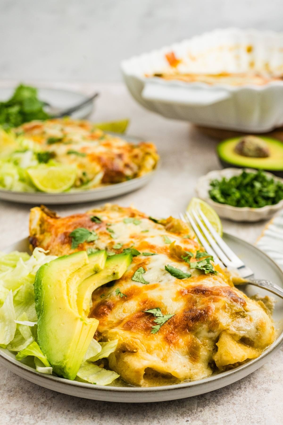 A plate of cheesy enchiladas served with sliced avocado, lettuce, and fresh cilantro on the side.