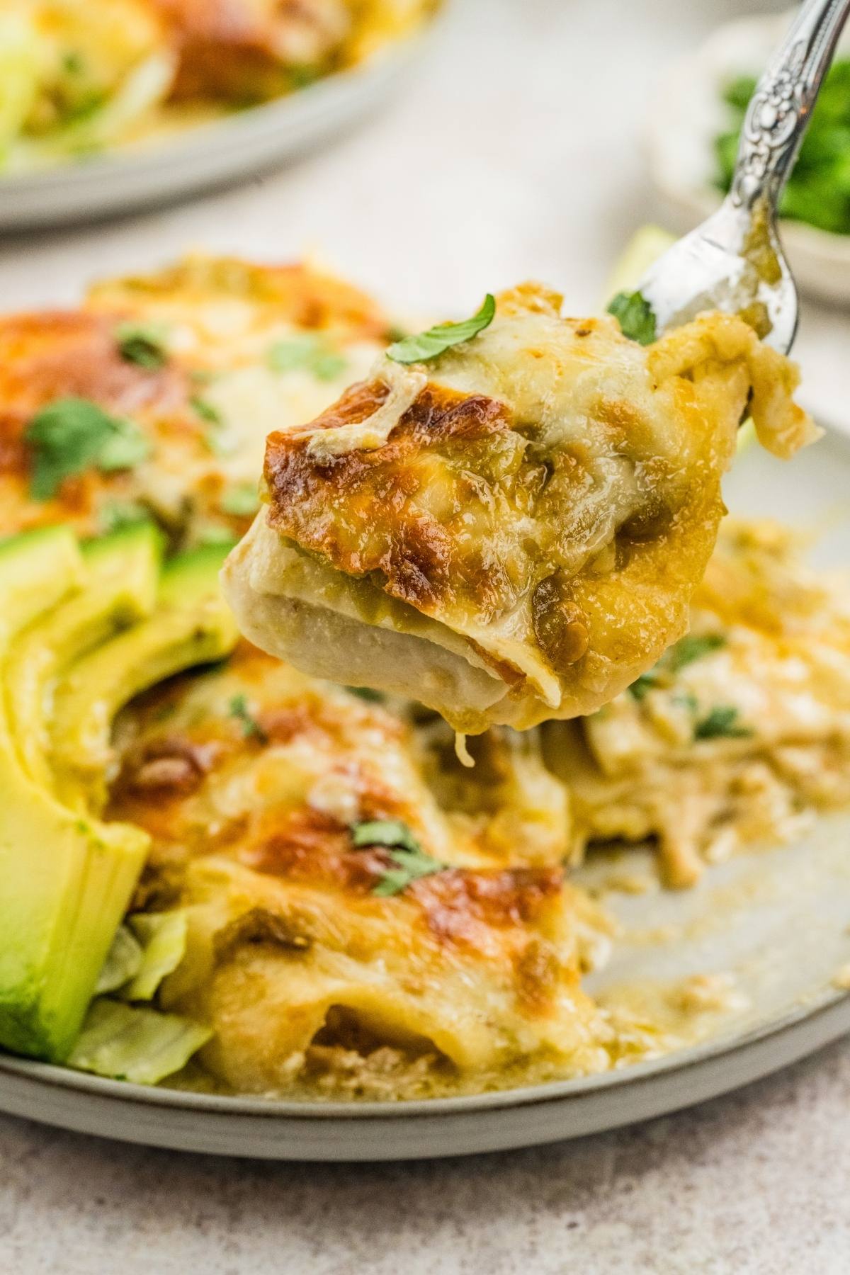 A fork holds a cheesy enchilada above a plate with avocado slices and more enchiladas.