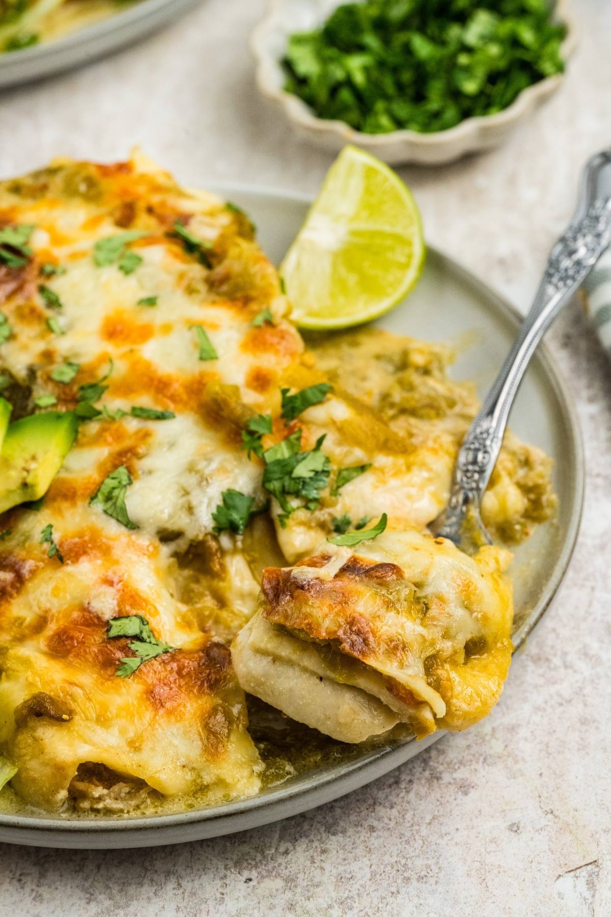 A plate of cheesy enchiladas topped with cilantro, served with a lime wedge and a fork.