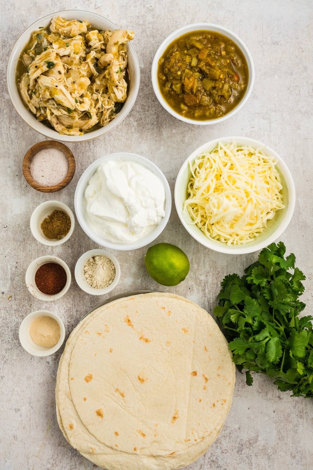 Ingredients for enchiladas: shredded chicken, tortillas, cheese, seasonings, sour cream, salsa, lime, cilantro.