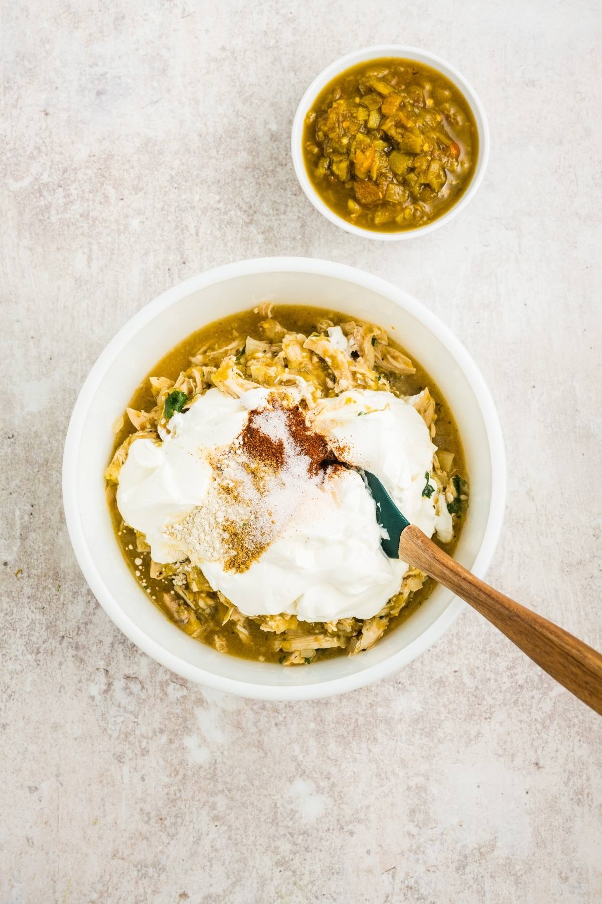 White bowl with shredded chicken, green sauce, sour cream, and spices; small bowl of green salsa nearby.