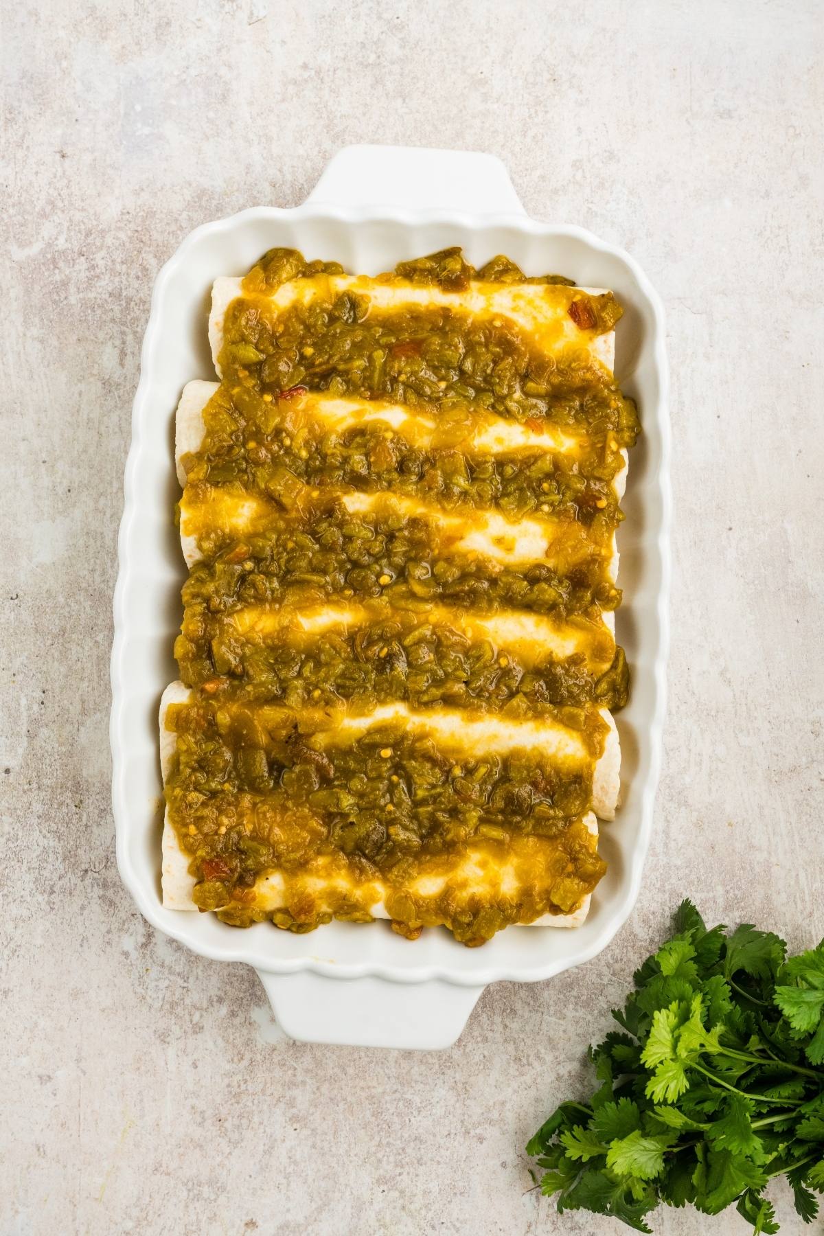 White casserole dish with enchiladas covered in green sauce, next to a bunch of fresh cilantro.