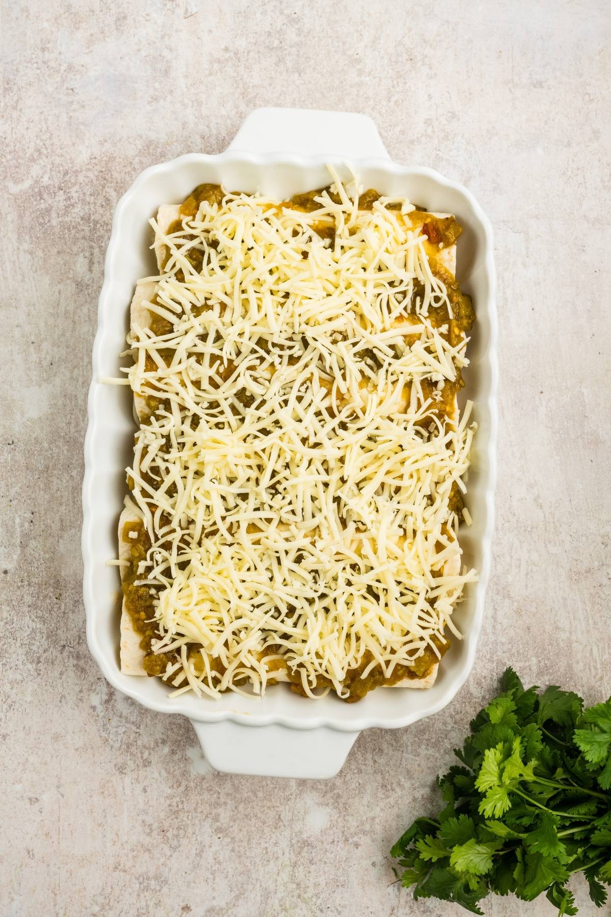 A white baking dish filled with enchiladas topped with shredded cheese, next to a bunch of fresh cilantro.