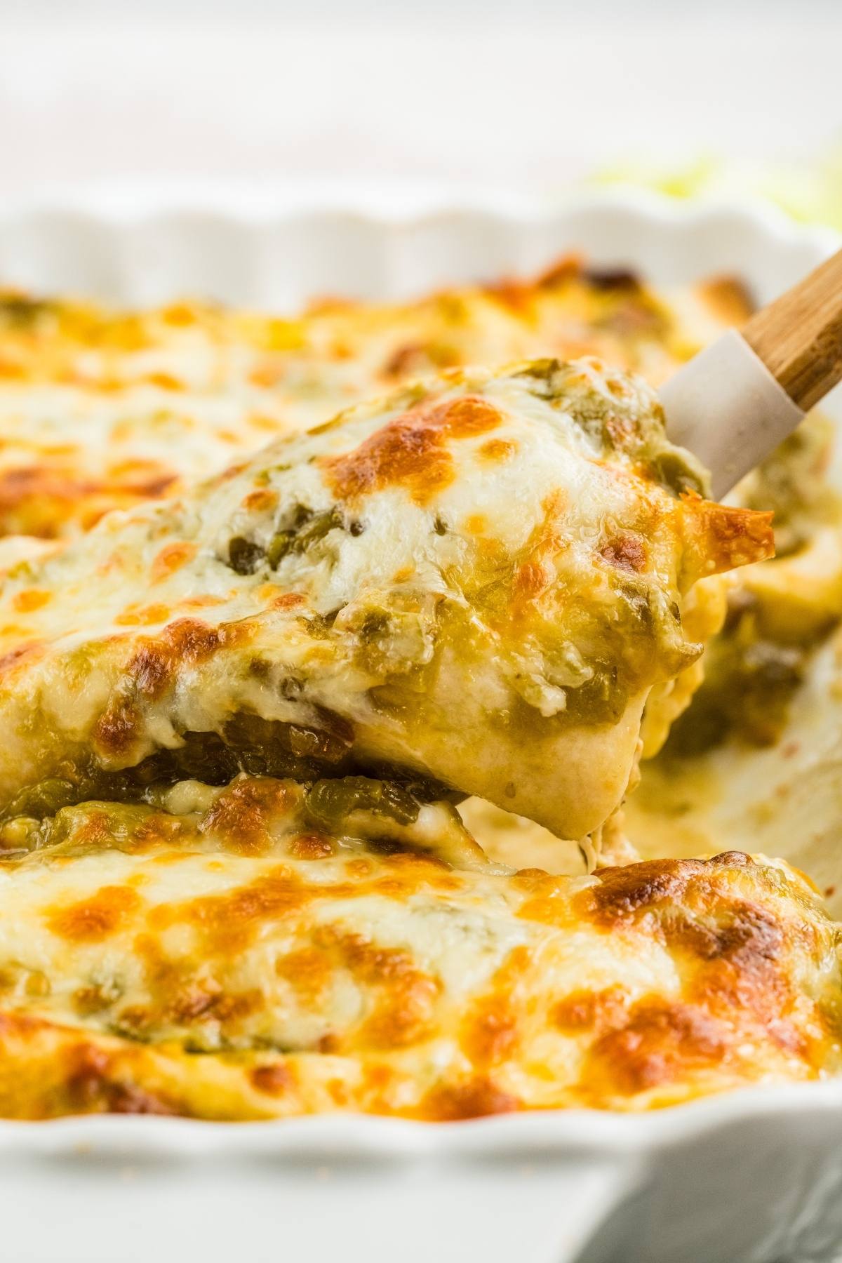 A spatula lifts a cheesy, golden-brown slice of baked enchiladas from a casserole dish.
