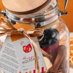 A Sangria Kit Gift Jar with a wooden lid, holiday tag, and ribbon is filled with festive items and sits next to dried orange slices.