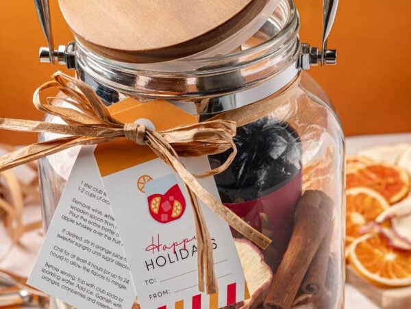 A Sangria Kit Gift Jar with a wooden lid, holiday tag, and ribbon is filled with festive items and sits next to dried orange slices.