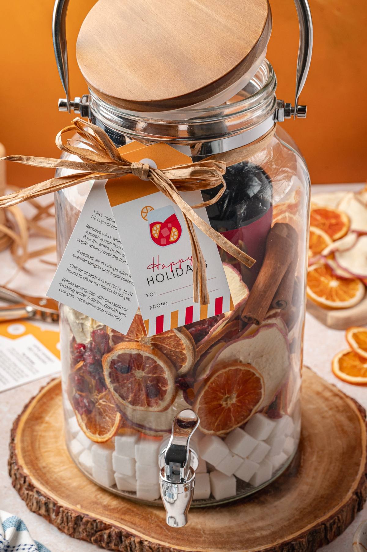 A glass jar with dried citrus slices, sugar cubes, and a holiday gift tag sits on a wooden slab.