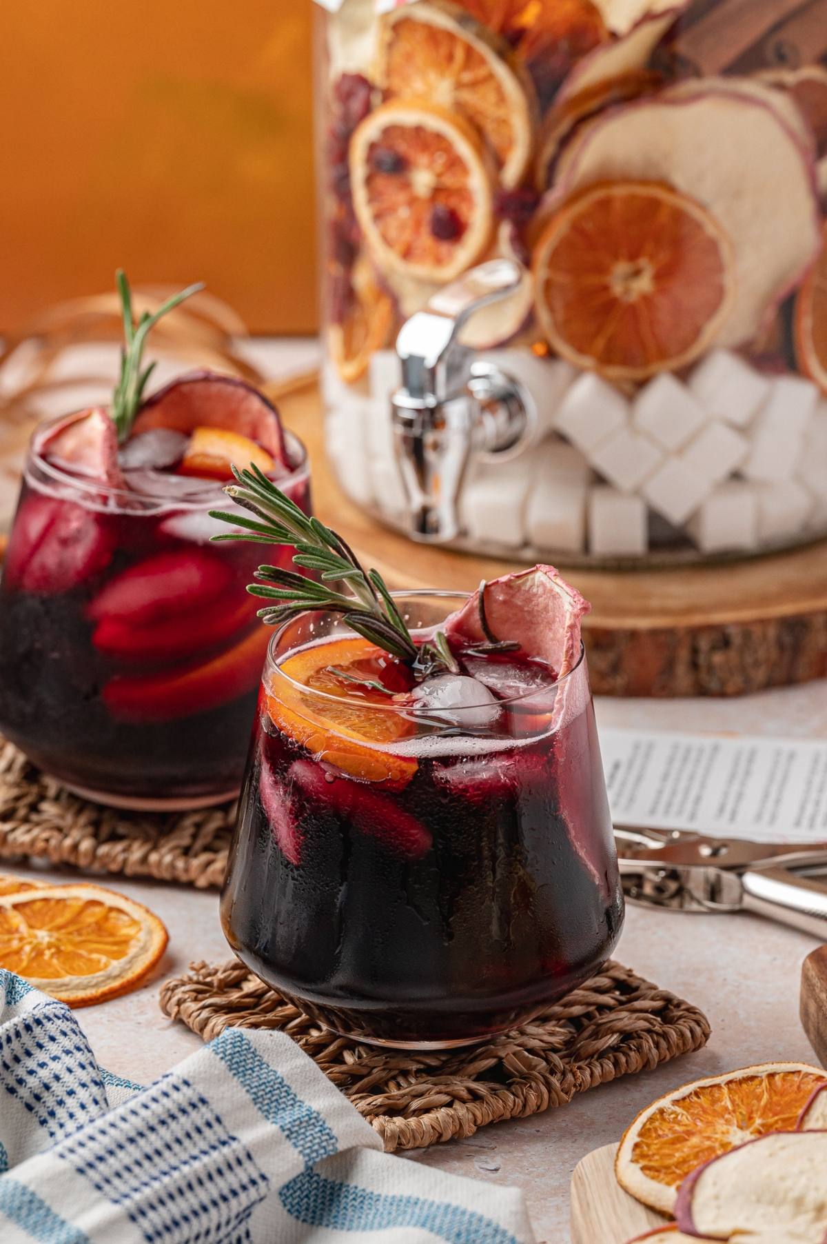 Two glasses of iced sangria with orange slices and rosemary garnish, next to a drink dispenser with dried citrus.