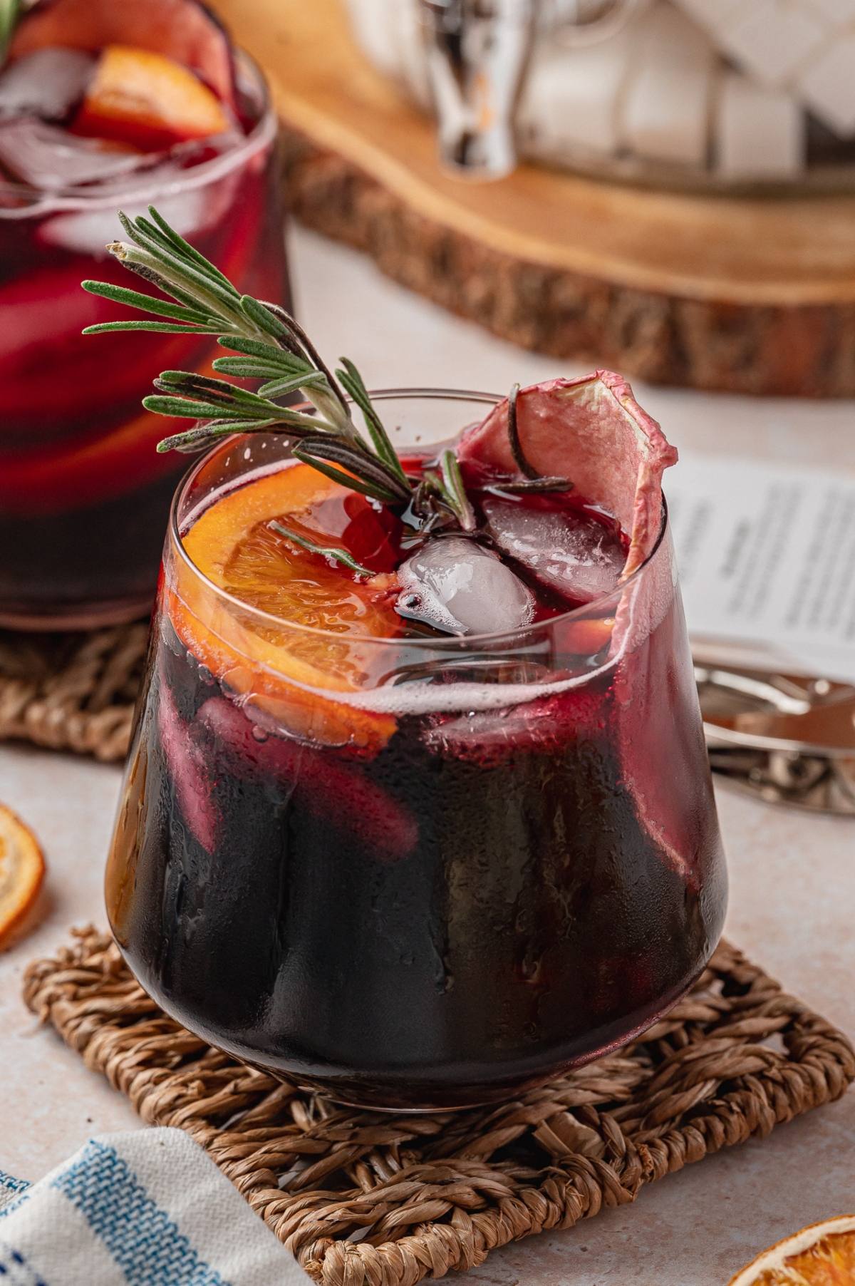 A glass of iced red sangria garnished with orange slices and a sprig of rosemary on a woven coaster.