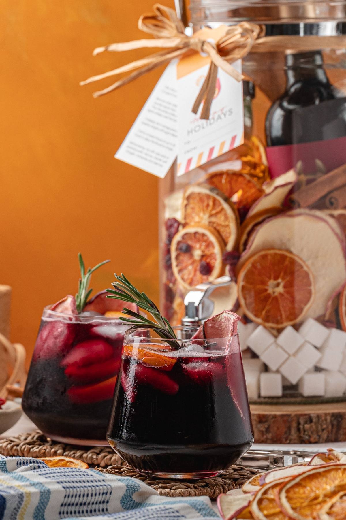 Two glasses of red drink with ice, orange slices, and rosemary, in front of a jar with dried citrus fruits.