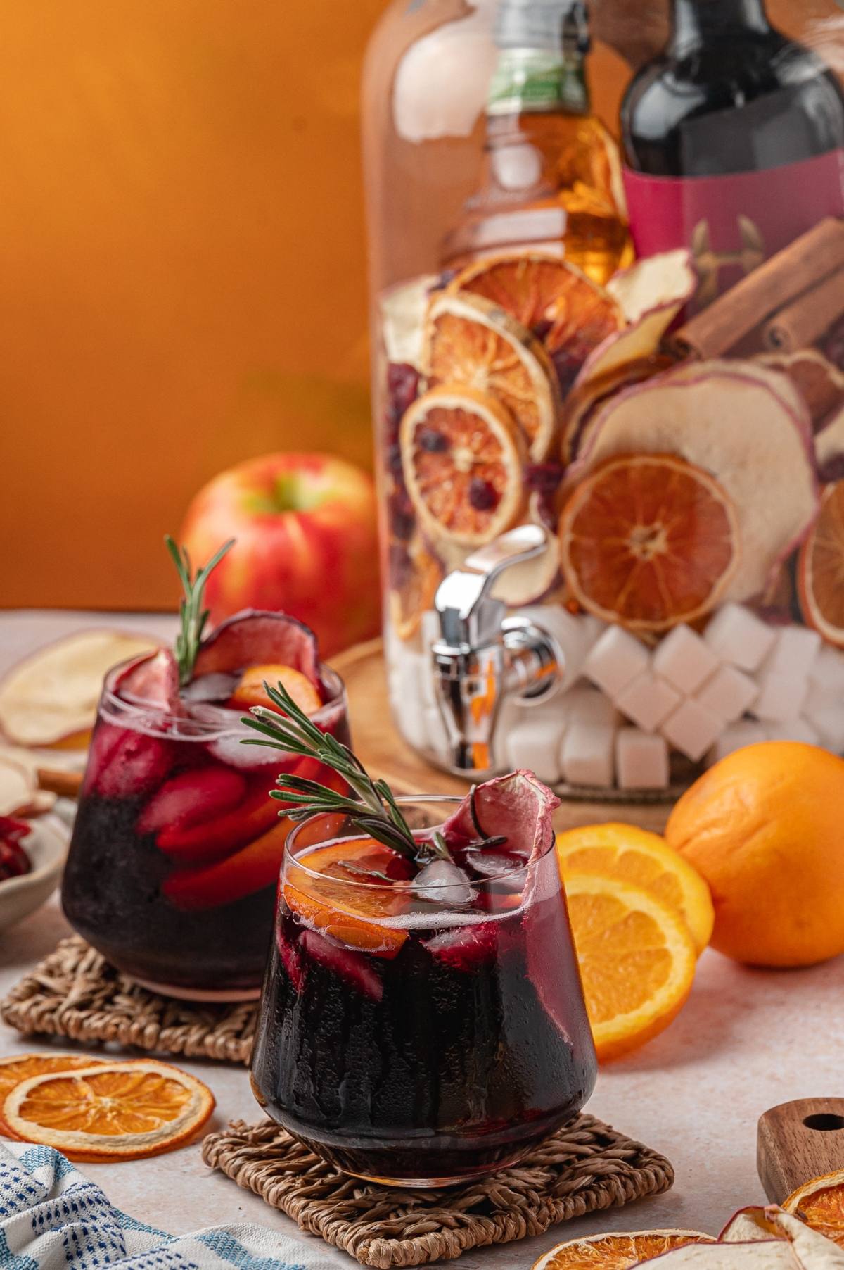 Two glasses of sangria with ice, orange slices, and rosemary, in front of a drink dispenser with dried fruit.