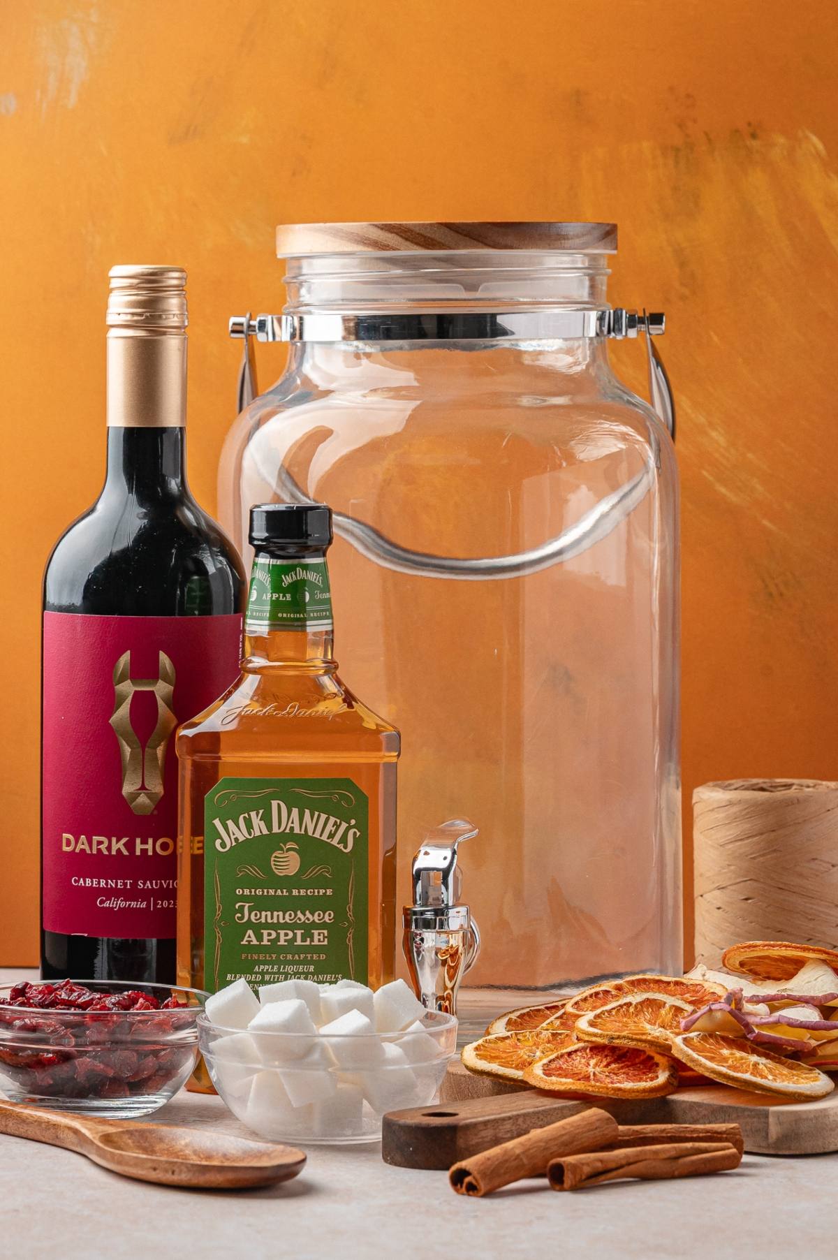 A bottle of red wine, Jack Daniel's whiskey, an empty glass jar, dried oranges, marshmallows, and spices.