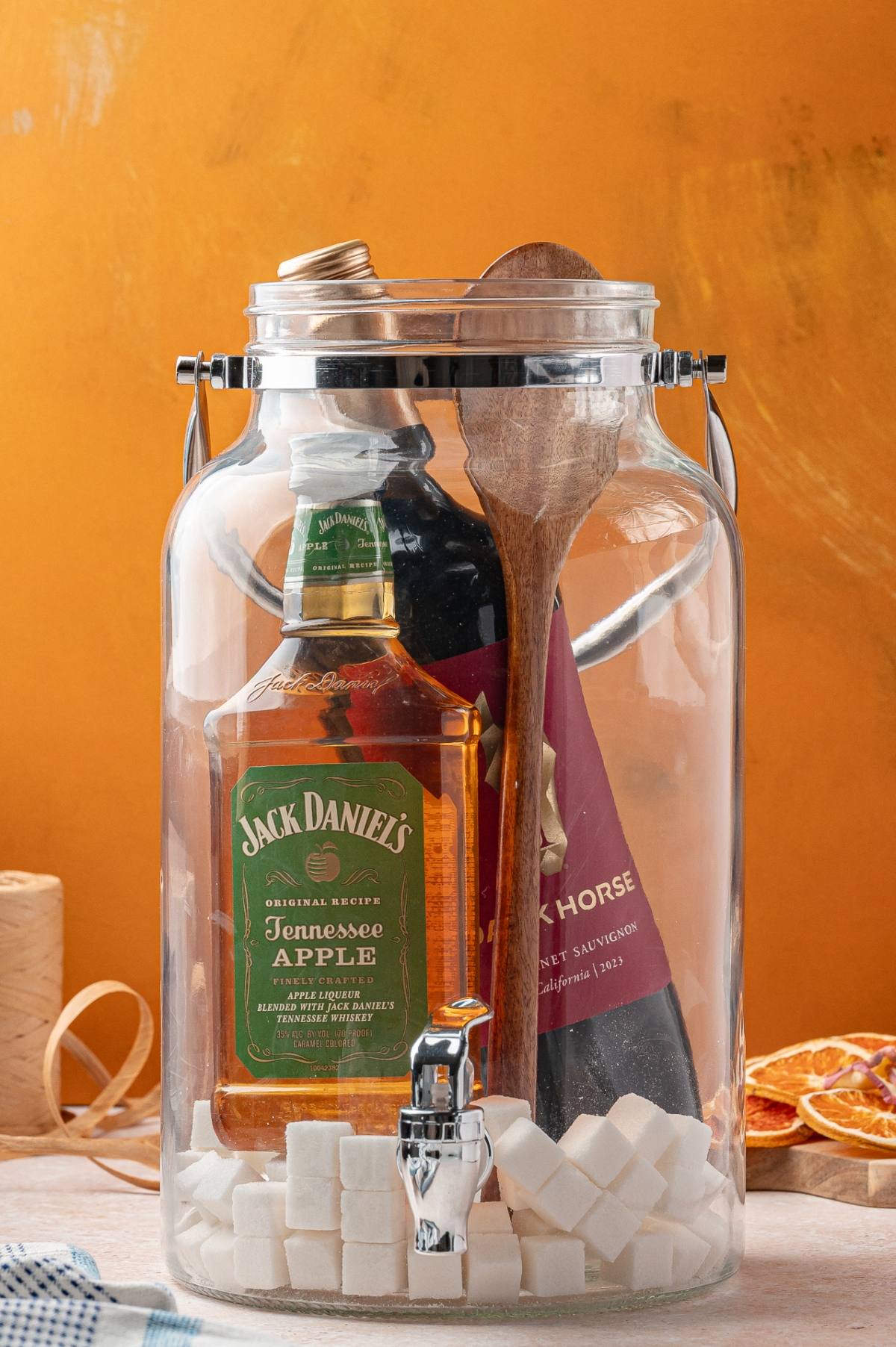 A glass dispenser with Jack Daniel’s Apple, red wine, wooden spoons, and sugar cubes against an orange background.