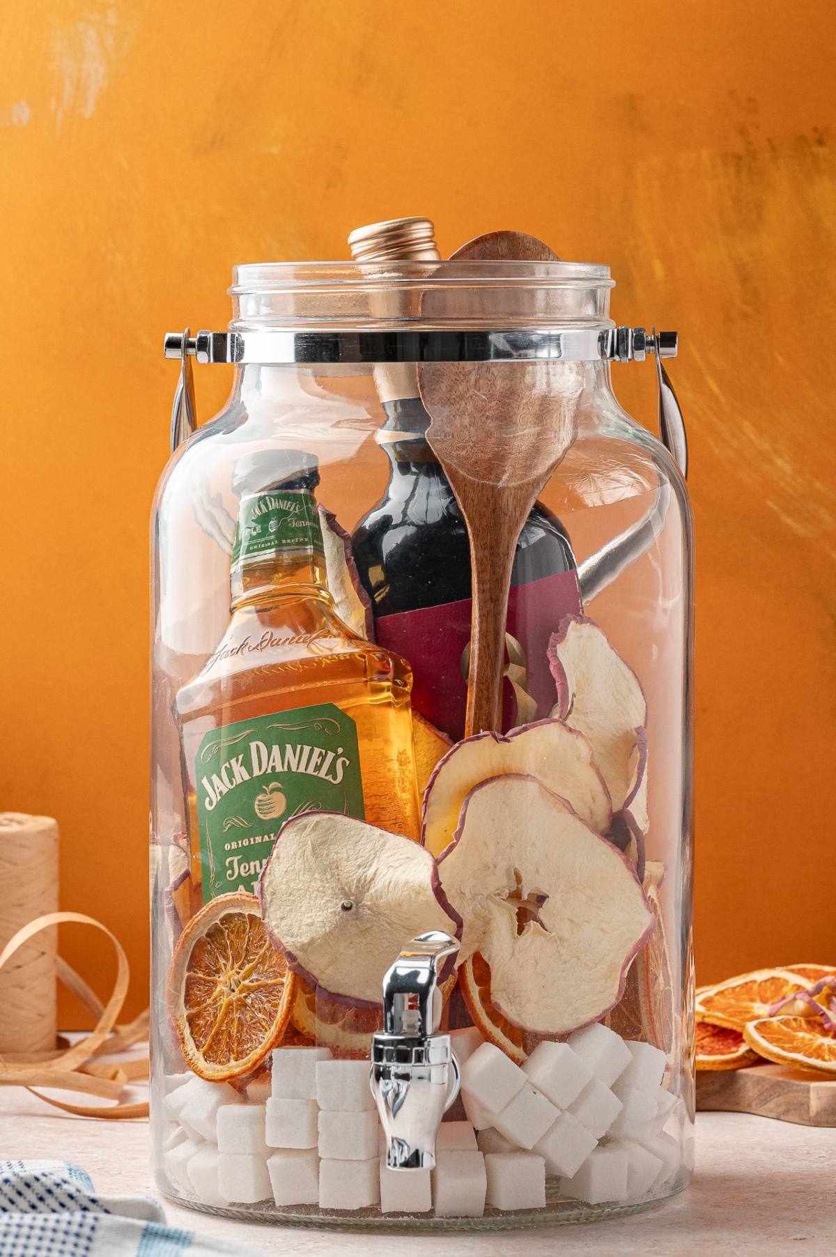 A jar filled with bottles, dried fruit, sugar cubes, and wooden utensils against an orange background.