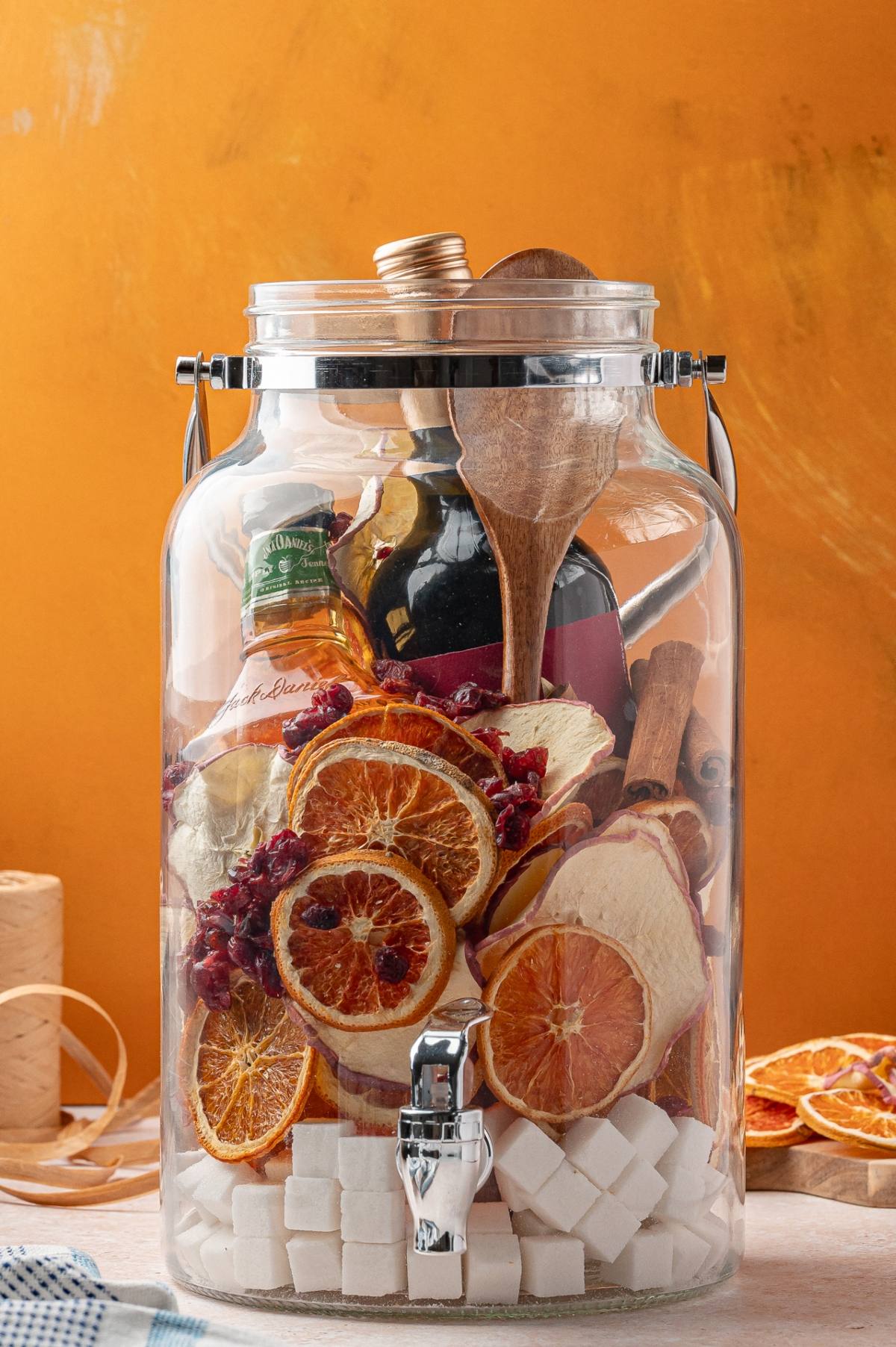 A glass jar with bottles, dried fruits, wooden spoons, cinnamon sticks, and sugar cubes against an orange background.