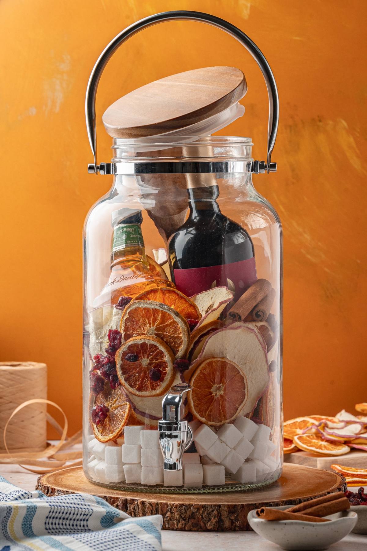 A glass drink dispenser filled with bottles, dried citrus slices, spices, and sugar cubes against an orange background.