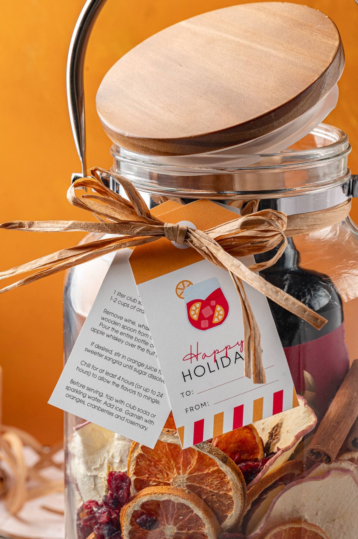 Glass jar with dried citrus slices, a cinnamon stick, gift tag, and wooden lid against an orange background.