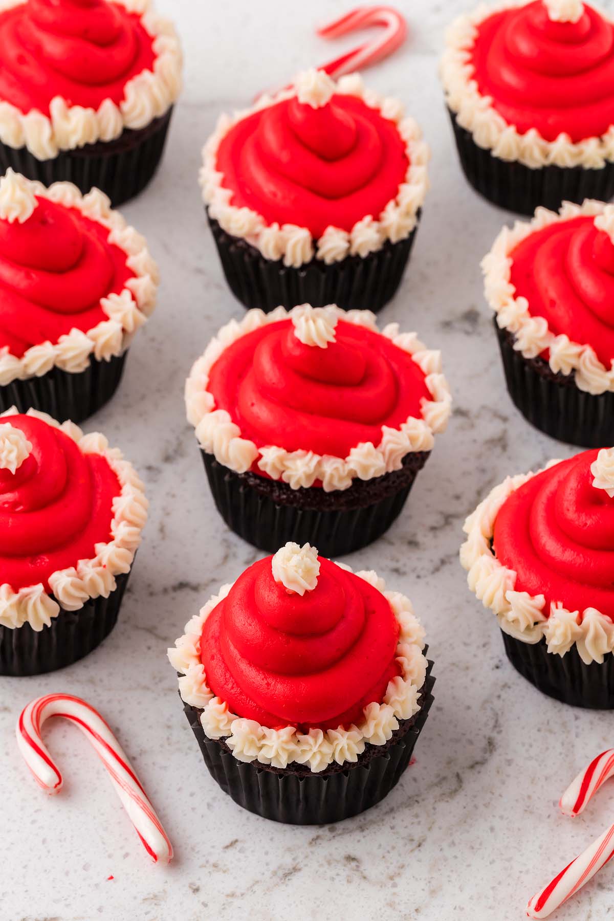 Nine Santa Hat Cupcakes with red and white frosting are arranged on a white surface, with candy canes nearby.