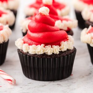 Santa Hat Cupcakes: Chocolate cupcakes with red and white icing shaped like Santa hats, topped with candy canes on a white surface.