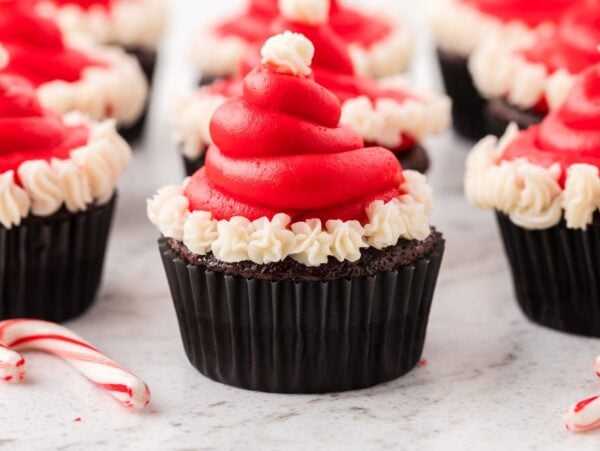 Santa Hat Cupcakes: Chocolate cupcakes with red and white icing shaped like Santa hats, topped with candy canes on a white surface.