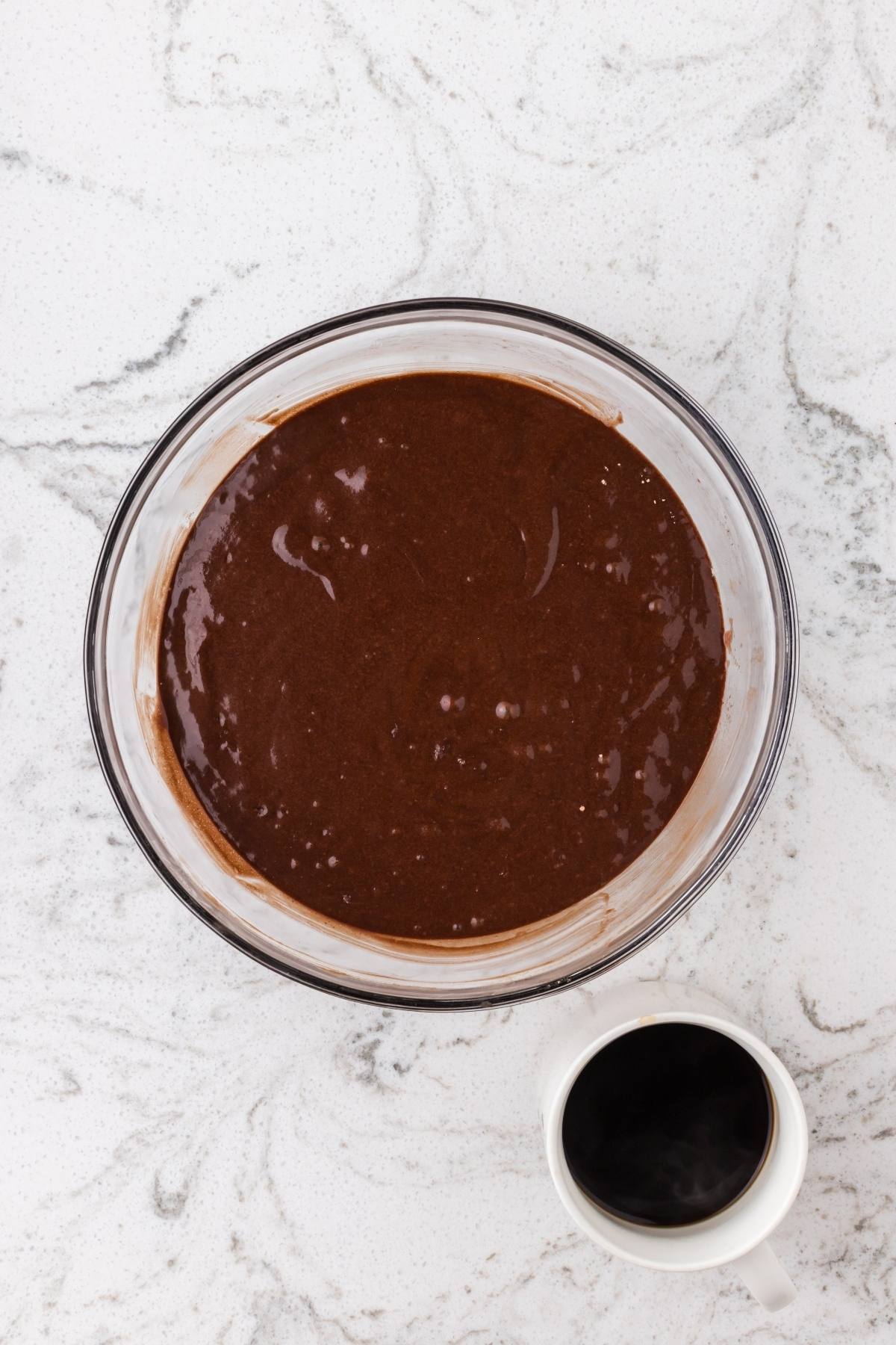 A glass bowl of chocolate cake batter next to a white cup of black coffee on a marble surface.
