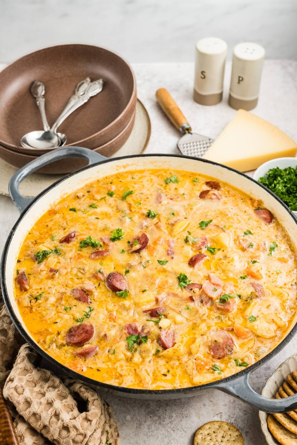 A pot of creamy sausage and potato chowder garnished with herbs, surrounded by bowls, cheese, and crackers.