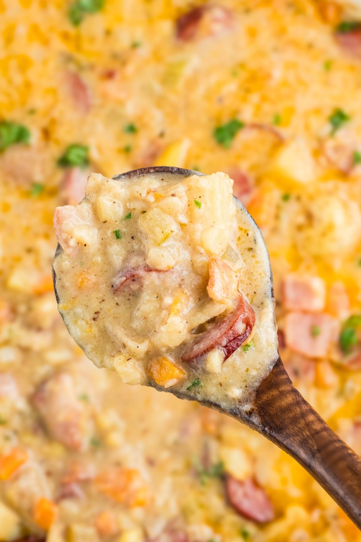 A wooden spoon holds creamy potato soup with chunks of sausage, potatoes, and herbs over a pot of soup.