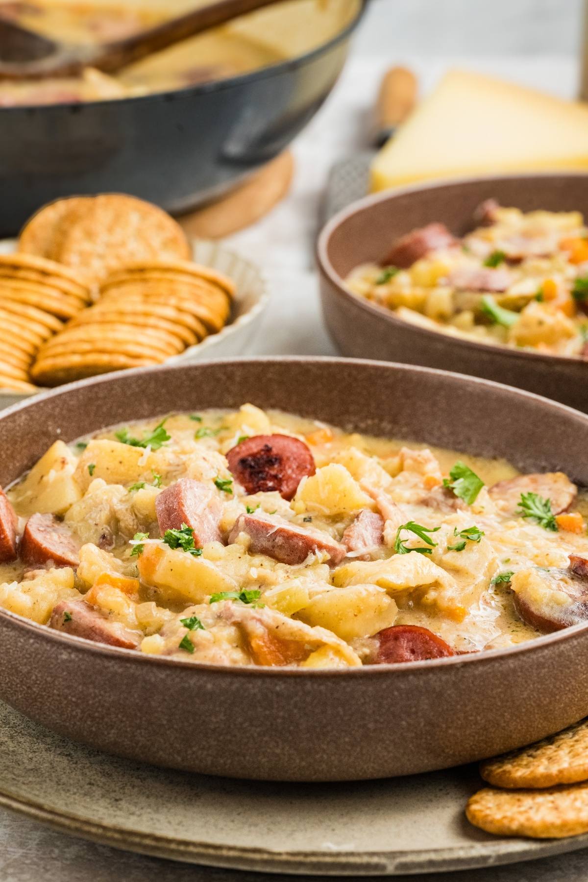 A bowl of hearty stew with sausage, vegetables, and crackers on the side.