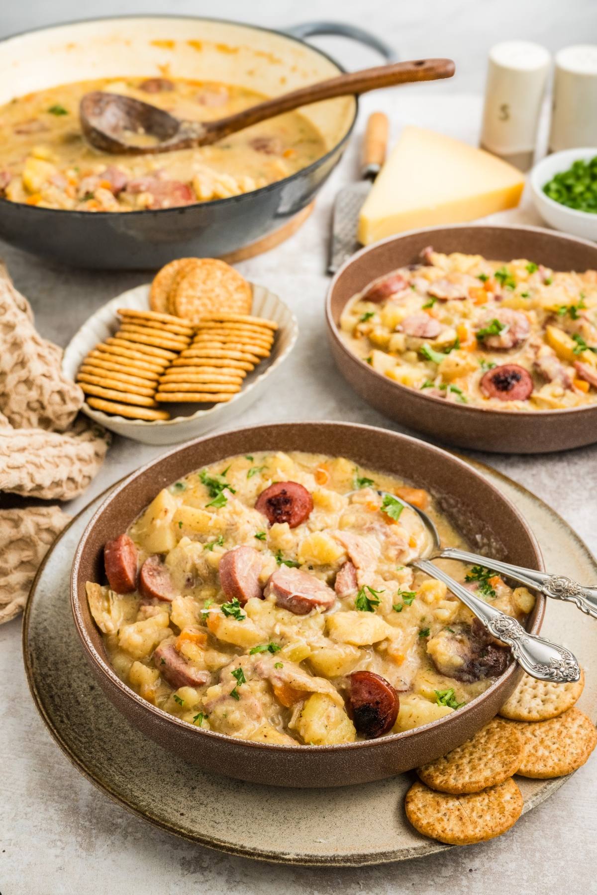 Two bowls of creamy sausage and potato soup with crackers, and a pot of soup in the background on a table.