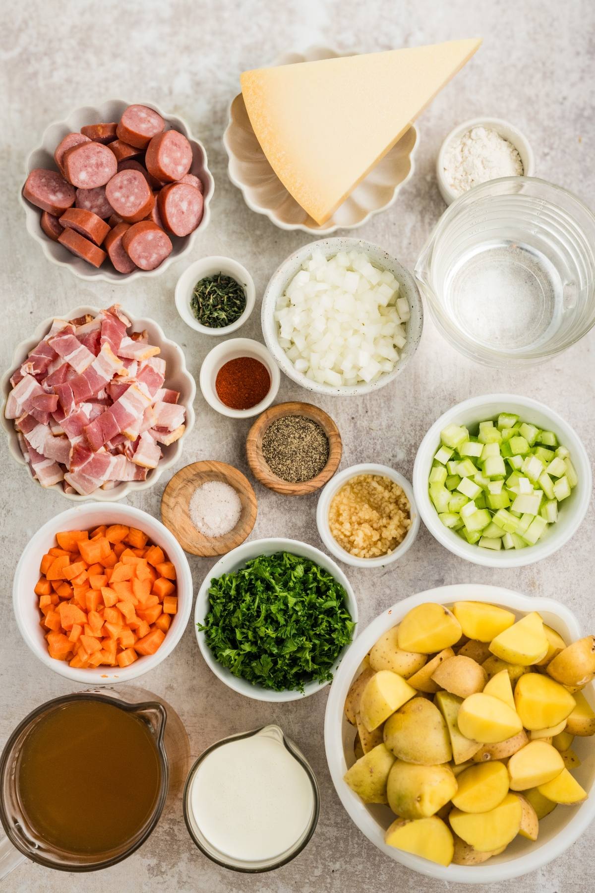Chopped sausage, bacon, veggies, herbs, cheese, broth, and cream arranged in bowls on a light surface.