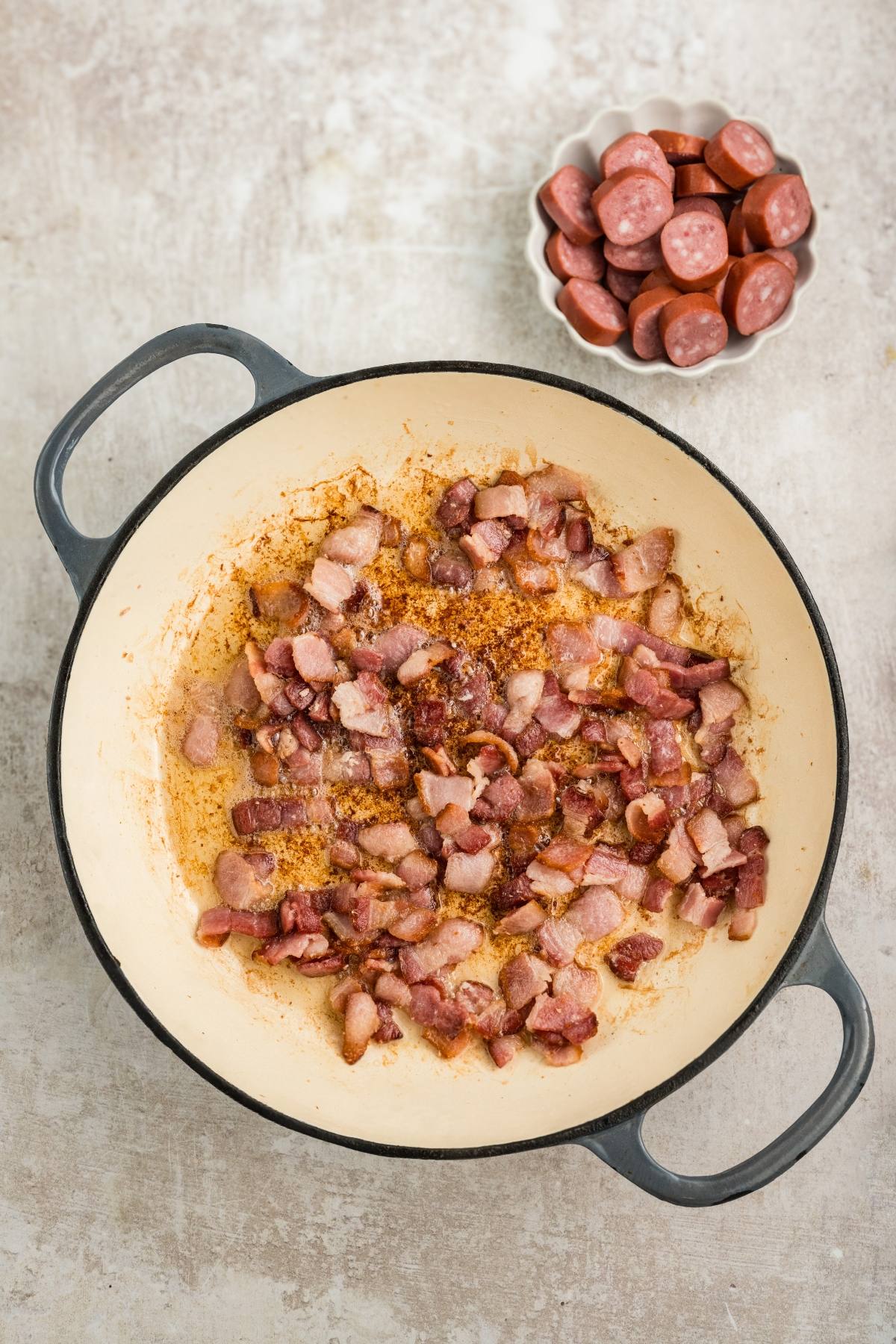 Chopped bacon cooking in a large pot, with sliced sausage in a bowl nearby.
