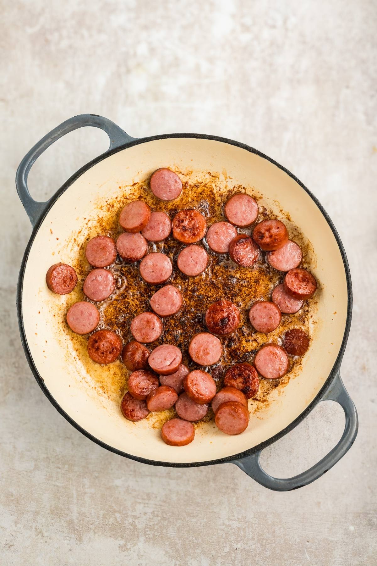 Sliced sausage browning in a large white pot with browned bits on the bottom.