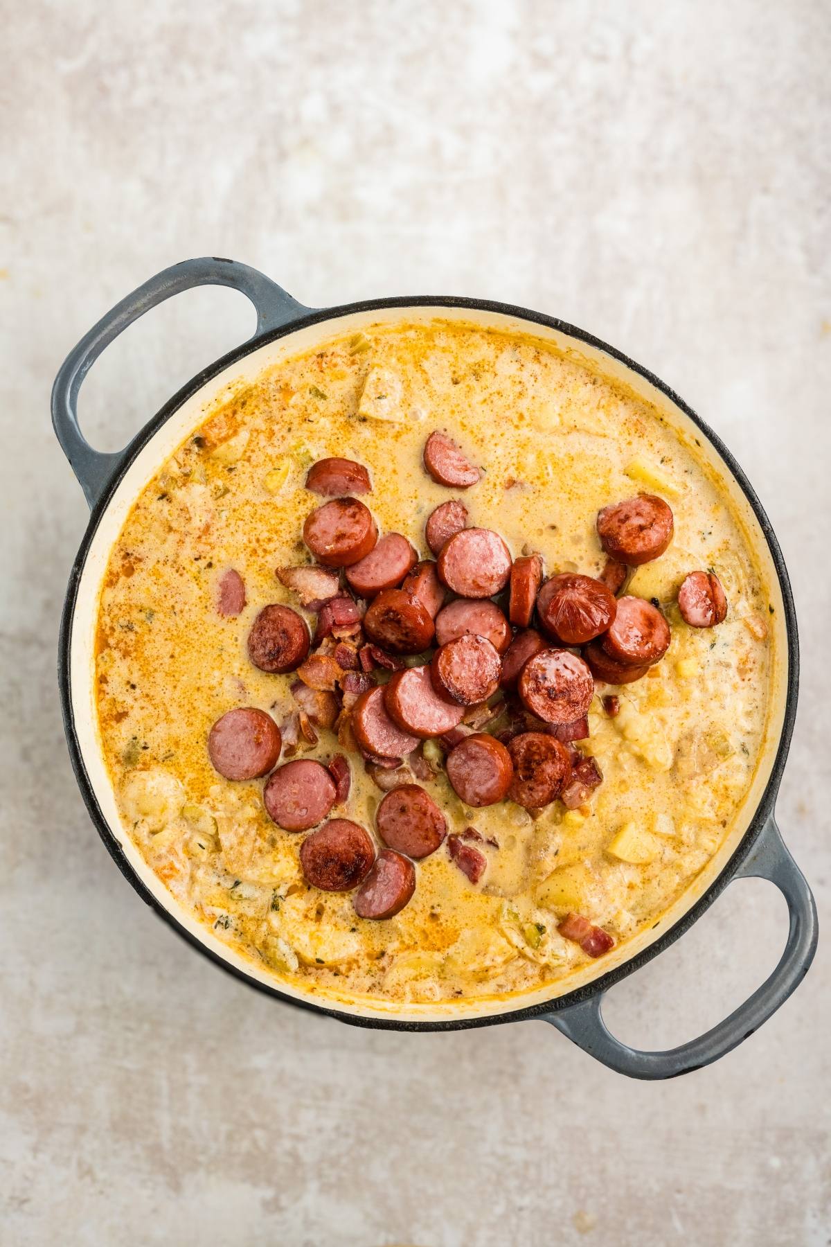 A pot of creamy soup topped with sliced, browned sausage on a light surface.