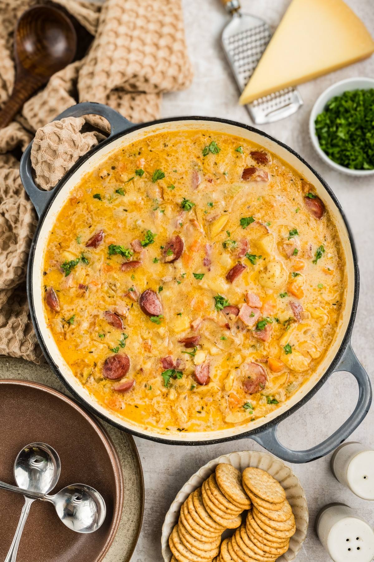 Creamy sausage and potato soup garnished with herbs in a pot, surrounded by crackers and cheese.