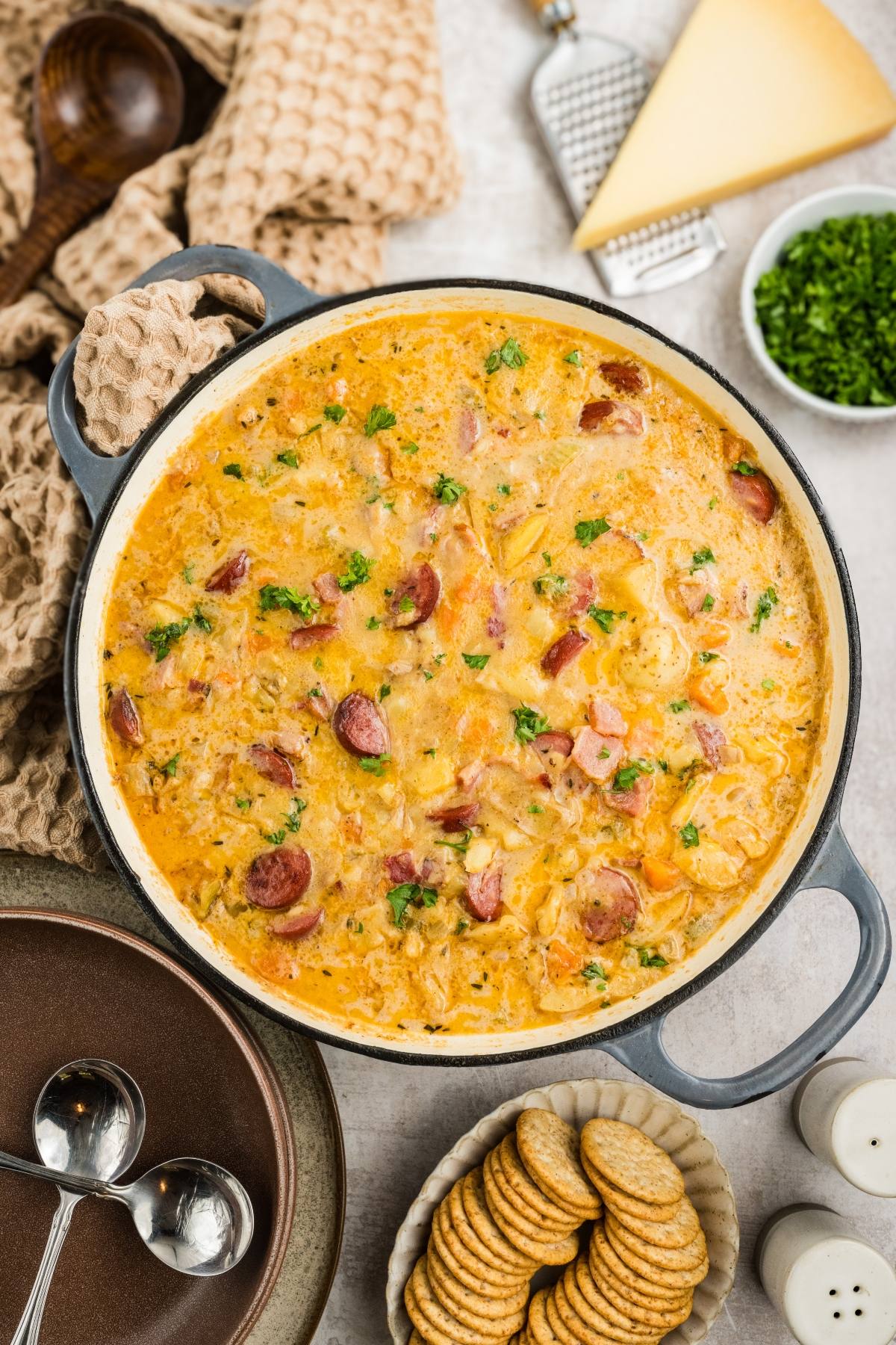 A pot of creamy sausage and potato soup garnished with herbs, with crackers and cheese on the side.